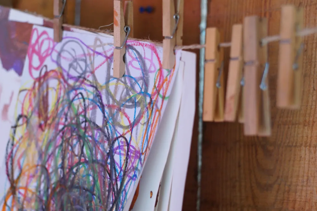Primer plano de varias láminas con garabatos de colores sujetas con pinzas de madera a un cordel, detalle del aula que muestra cómo el equipo expone y acompaña los procesos creativos de las niñas y los niños.