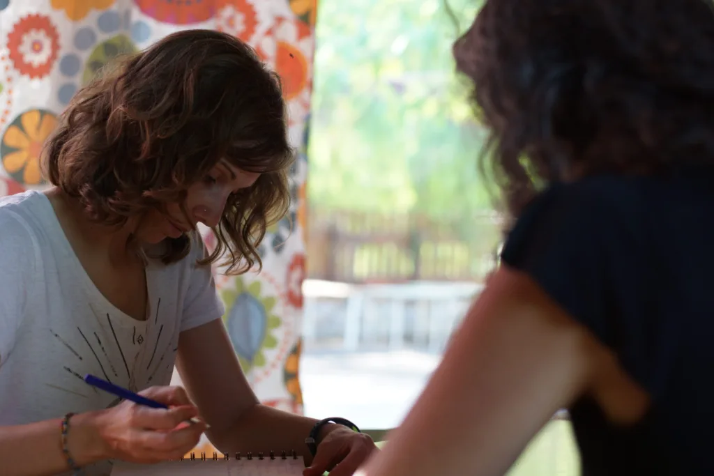 Integrante del equipo anota en un cuaderno durante una reunión de coordinación con otra compañera; se aprecia una cortina floral y luz natural del espacio educativo, transmitiendo trabajo colaborativo y cuidado.