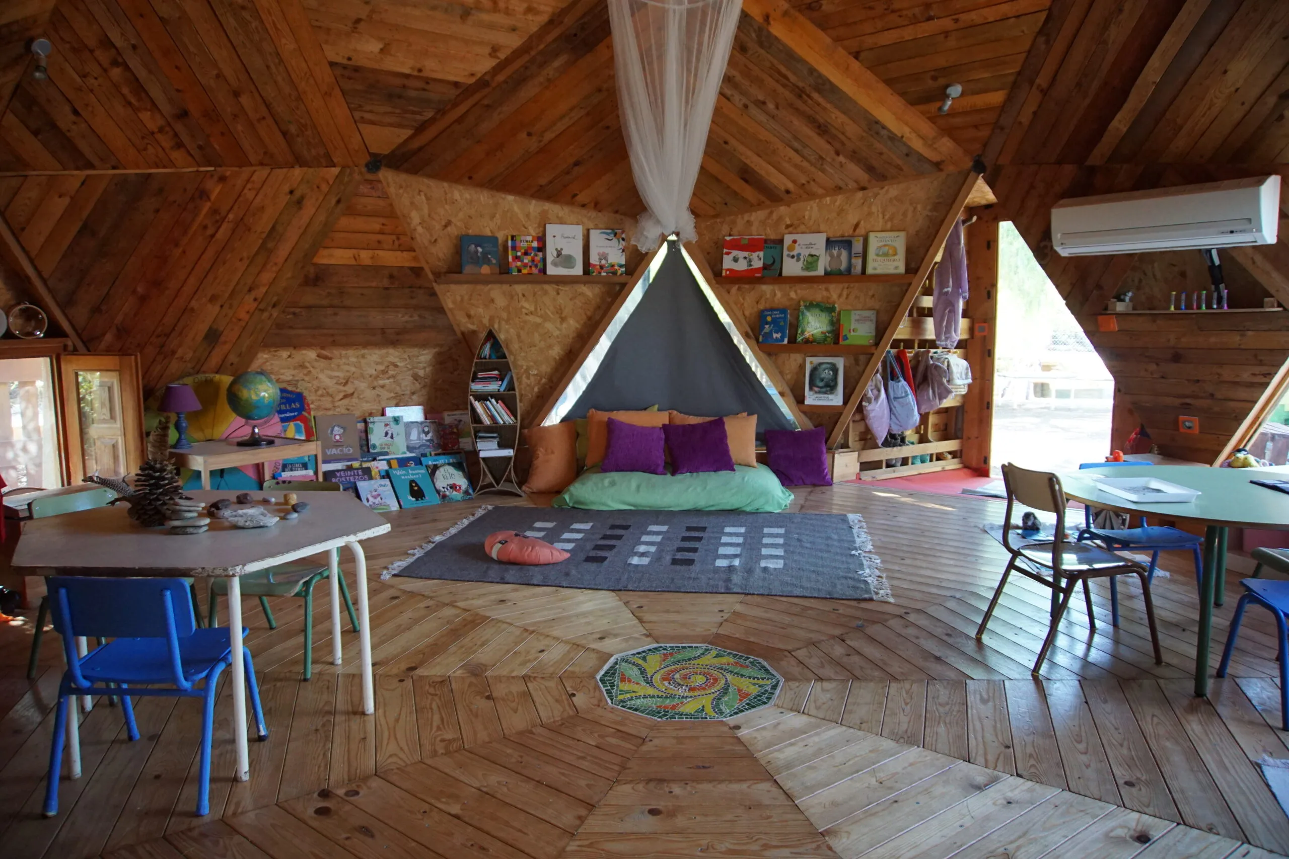 Aula en domo geodésico de madera con rincón de lectura, mesas infantiles y luz natural en un centro en la naturaleza.