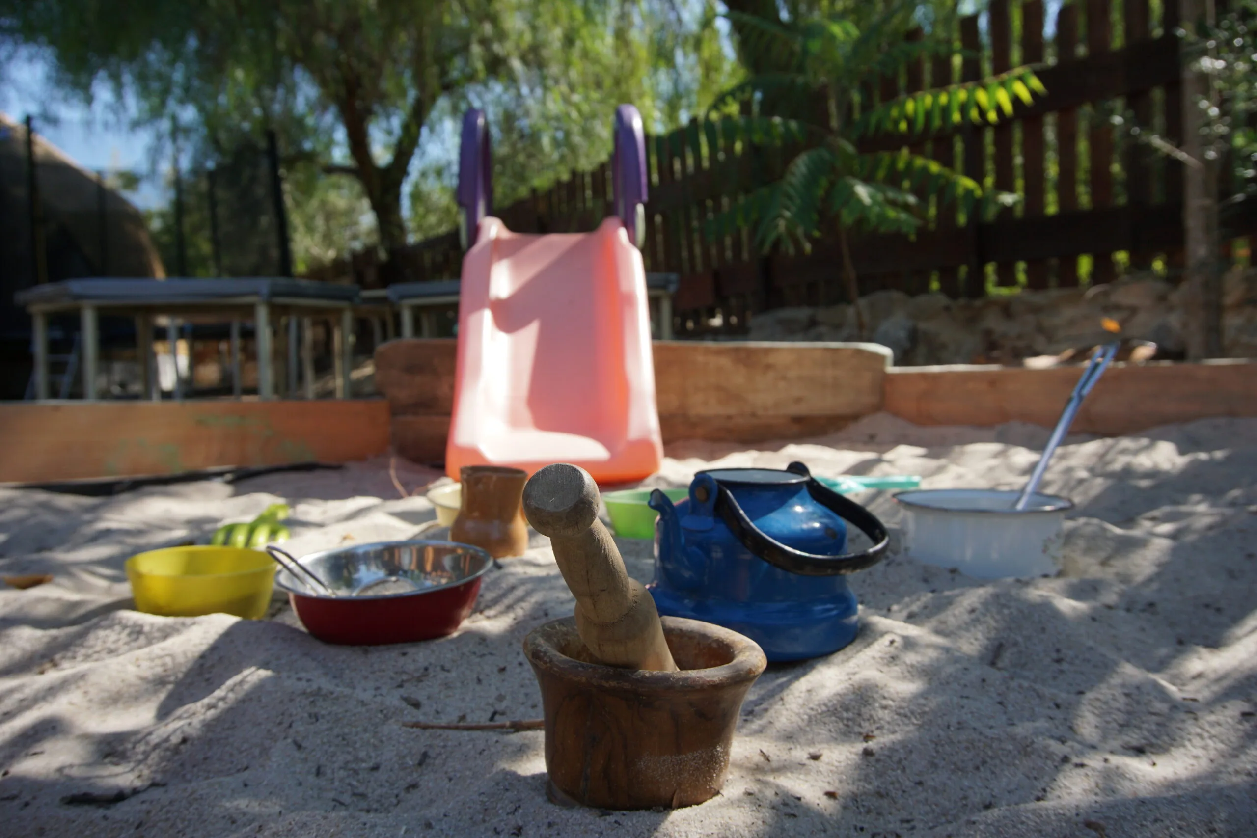 Utensilios de juego en arena (mortero de madera, tetera y cuencos) con tobogán exterior; aprendizaje libre y en contacto con la naturaleza en nuestro espacio educativo.
