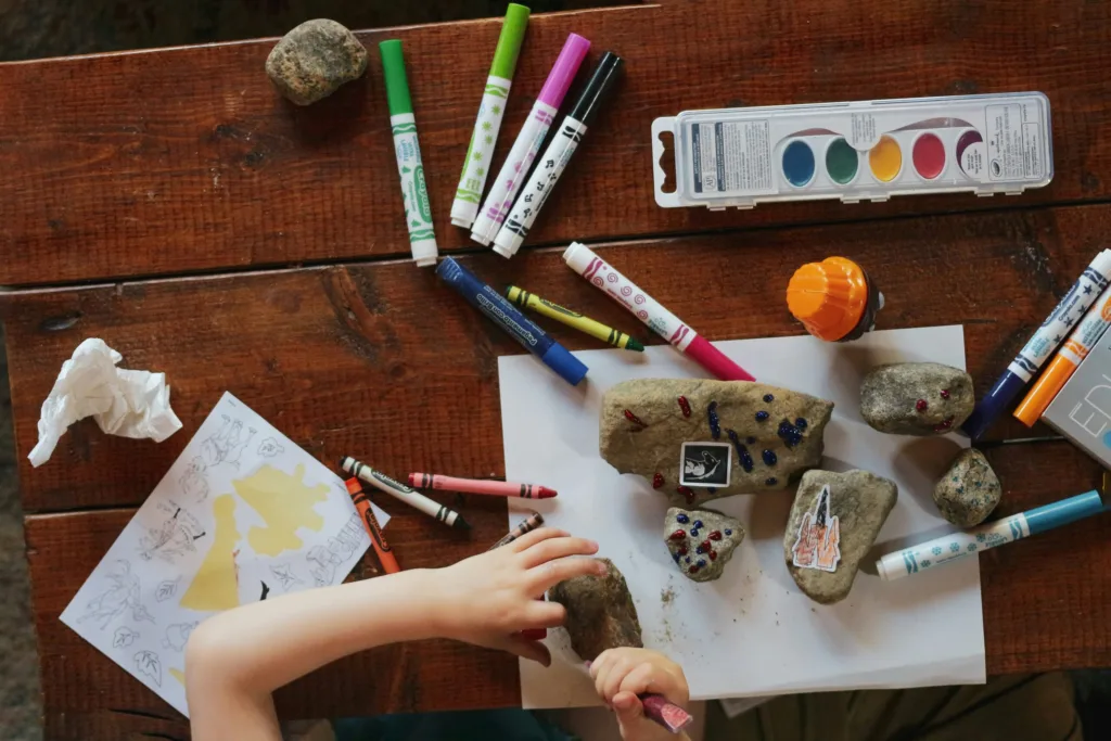 Manos infantiles decoran piedras con rotuladores y pegatinas sobre mesa de madera; actividad artística y motricidad fina en un espacio educativo.