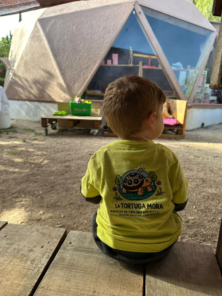 Niño pequeño de espaldas con camiseta de La Tortuga Mora frente a un domo geodésico usado como aula; aprendizaje en la naturaleza en un espacio educativo.