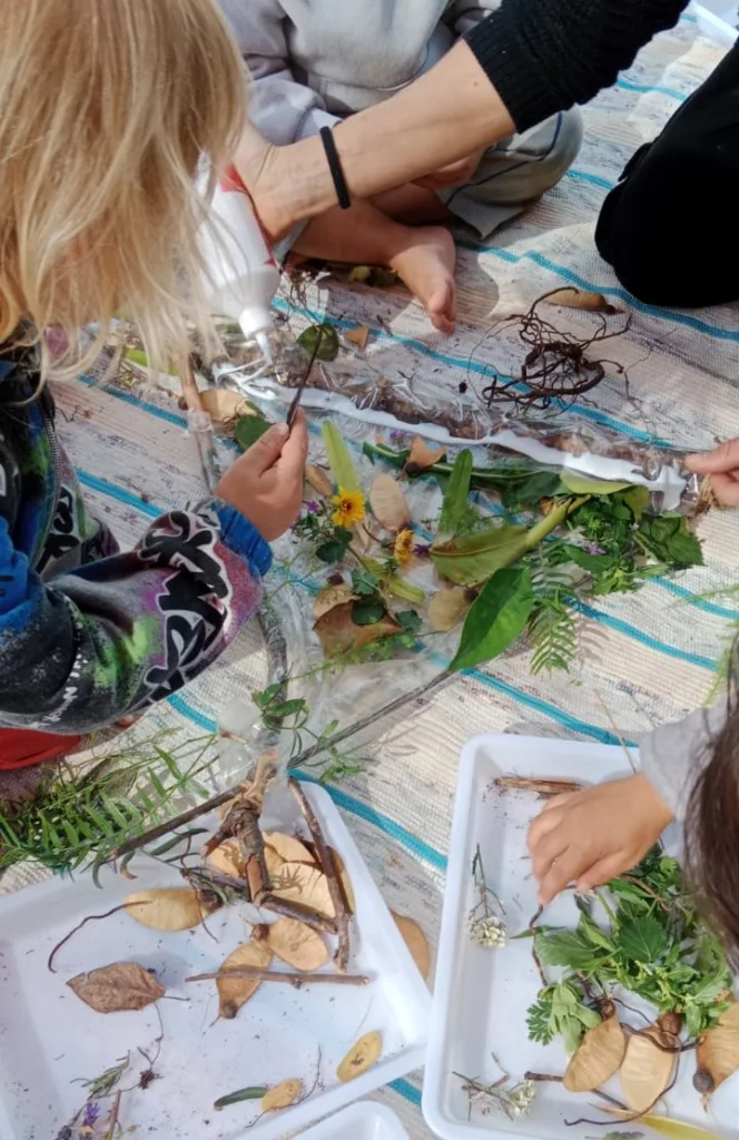 Niños elaborando un collage con hojas, flores y ramas al aire libre; aprendizaje sensorial y creatividad en nuestro espacio educativo.