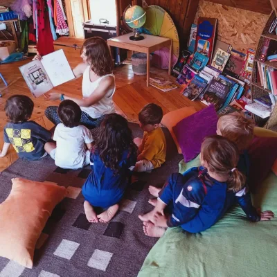 Actividades de cuentacuentos una educadora lee un libro a niños sentados en alfombra, aula con estanterías de libros y cojines.