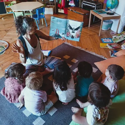 Actividades de cuentacuentos en aula de madera una educadora muestra un libro ilustrado a un grupo de niños sentados en la alfombra, con cojines y estanterías de libros alrededor.