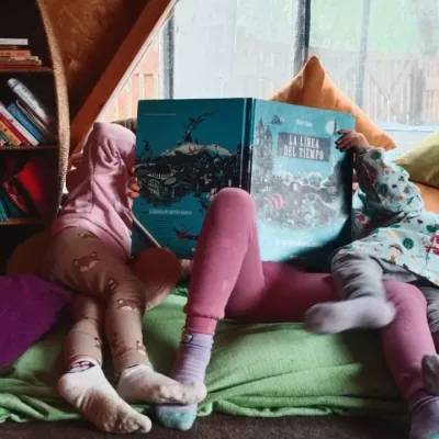 Actividades de lectura infantil, tres niños leen un libro grande en rincón con cojines y estanterías dentro de un domo geodésico rodeados de libros.