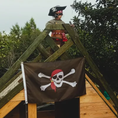 Actividades de juego simbólico un niño disfrazado de pirata sobre cabaña de madera con bandera corsaria; juego imaginativo al aire libre.