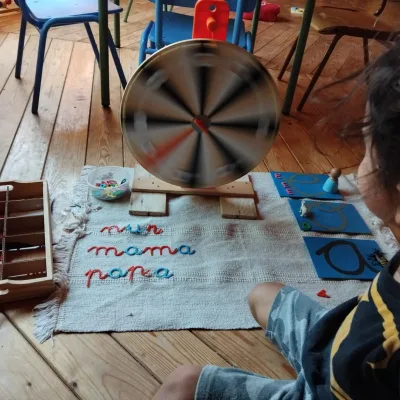 Actividades de lenguaje una niña usa alfabeto móvil sobre alfombra, forma palabras “mi, mamá, papá” y practica preescritura con apoyo de rueda giratoria.