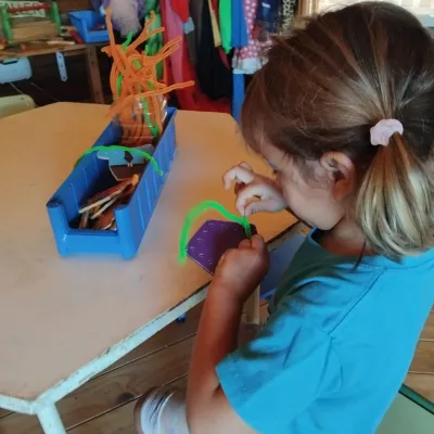 Actividades de motricidad fina una niña enhebra limpiapipas en un tablero perforado sobre mesa del aula; concentración y coordinación ojo-mano.