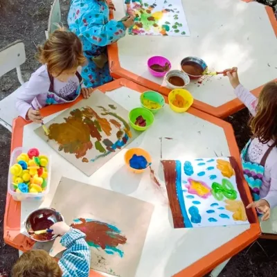 Actividades de arte al aire libre, niños pintan con pinceles y témperas en mesas hexagonales, explorando colores y trazos.