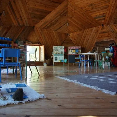 Aula en domo geodésico de madera con mesas y sillas azules, cocina de juego y perchero de disfraces; centro en la naturaleza.