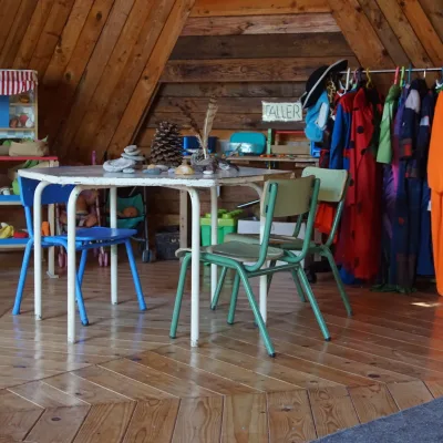 Mesa infantil con sillas, piedras y piñas para exploración; perchero de disfraces y puesto de mercado en aula de madera de un centro en la naturaleza.
