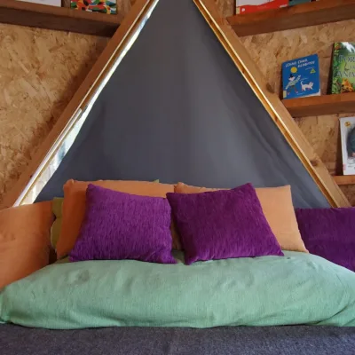 Rincón de lectura con colchoneta verde y cojines morados y naranjas bajo nicho triangular; estanterías de cuentos en aula de domo geodésico de un centro en la naturaleza.