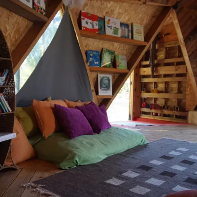 Rincón de lectura con colchoneta y cojines, estanterías triangulares y librería de mimbre junto a la entrada; aula de madera en un centro en la naturaleza.