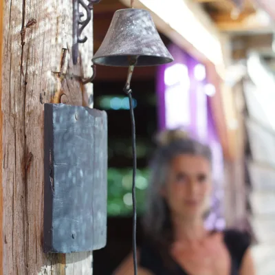 Primer plano de una campana metálica antigua colgada en un poste de madera con pequeña pizarra; al fondo, una persona desenfocada y telas moradas, detalle acogedor del centro en la naturaleza.