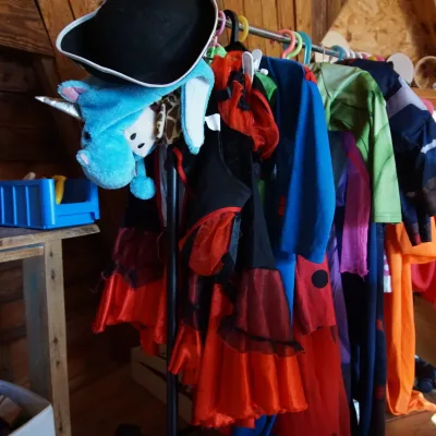 Primer plano de un perchero con disfraces infantiles de colores, sombrero y gorro de unicornio junto a una mesa de materiales, en una sala de madera del centro en la naturaleza, pensado para juego simbólico y creatividad.