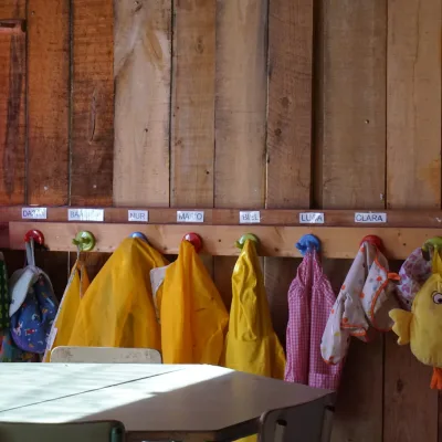Fila de percheros de colores con etiquetas de nombres, impermeables amarillos y babis colgados sobre pared de madera junto a mesas escolares; detalle de organización del aula en el centro en la naturaleza.