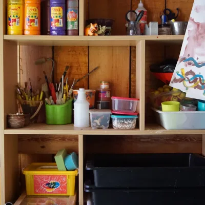 Estantería de madera con materiales de arte: botes de pintura, pinceles, tijeras, recipientes y bandejas, esponjas y cajas organizadoras junto a un dibujo infantil, detalle del taller creativo del centro en la naturaleza.