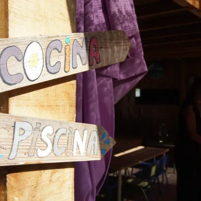 Carteles de madera pintados a mano con las palabras “Cocina” y “Piscina” en un porche de madera con cortinas moradas y mesas infantiles al fondo, detalle acogedor del centro en la naturaleza.