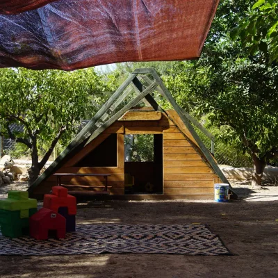Cabaña de madera con estructura tipo rocódromo en forma de A bajo malla de sombra, rodeada de árboles y huerto; en primer plano alfombra exterior con bloques blandos de colores y banco infantil, espacio de juego del centro en la naturaleza.
