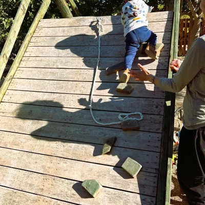 Niño pequeño subiendo una rampa de madera con presas y cuerda, acompañado por un adulto que le ofrece apoyo; actividad de psicomotricidad y equilibrio en la zona cabaña-rocódromo del centro en la naturaleza, bajo árboles y a cielo abierto.
