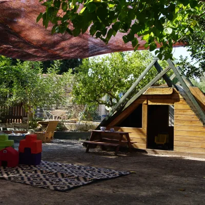 Cabaña infantil de madera con estructura tipo rocódromo bajo malla de sombra, alfombra con bloques blandos, mesa de picnic y tobogán al fondo entre árboles; área de juego del centro en la naturaleza.