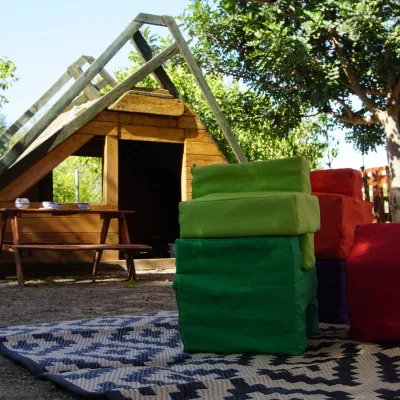 Primer plano de bloques blandos de colores sobre alfombra frente a una cabaña de madera con estructura tipo rocódromo y mesa de picnic, rodeada de árboles y valla; área de juego al aire libre del centro en la naturaleza.