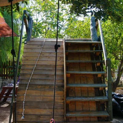 Estructura de juego exterior con rampa de madera tipo rocódromo y cuerda, escaleras laterales y columpio bajo la sombra de árboles; zona cabaña-rocódromo del centro en la naturaleza.