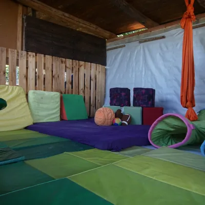Zona dinámica cubierta del centro en la naturaleza con colchonetas verdes, columpio de cuerda, tela aérea naranja, túneles sensoriales, cojines y pelotas; espacio seguro para psicomotricidad y juego libre infantil.