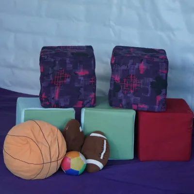Primer plano de la zona dinámica del centro en la naturaleza con cubos acolchados de colores, cojines morados y pelotas blandas (baloncesto, rugby y multicolor) sobre colchoneta violeta, material pensado para juego sensorial y desarrollo de la motricidad gruesa infantil.