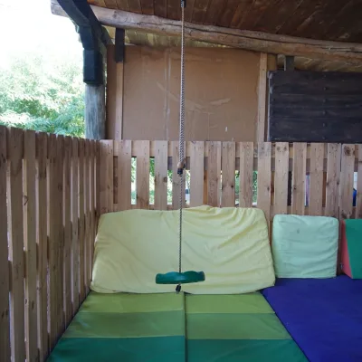 Zona dinámica del centro en la naturaleza: columpio de cuerda con asiento verde colgado del techo de madera, suelo con colchonetas verdes y base morada, grandes cojines amarillos como respaldo y cerramiento de lamas de madera; espacio cubierto y seguro para juego libre y motricidad gruesa infantil.