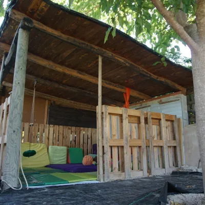 Porche de madera con paredes de palets, colchonetas y cojines para psicomotricidad y juego libre; zona dinámica a la sombra de un árbol en el centro en la naturaleza.