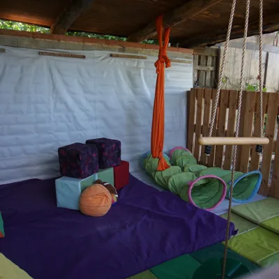 Espacio de zona dinámica del centro en la naturaleza: área cubierta con colchonetas verdes y base morada, tela aérea naranja, columpio de barra de madera con cuerdas, túneles sensoriales verdes y cubos blandos; rincón seguro para juego libre y motricidad gruesa infantil.