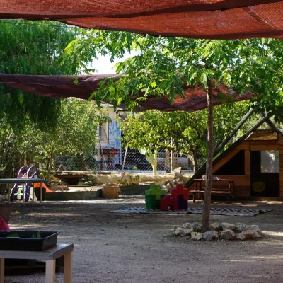 Patio con toldos de sombra, cabaña de madera tipo casita, mesa de picnic, bloques de espuma y arenero con tobogán entre árboles; zona exterior de un centro en la naturaleza.
