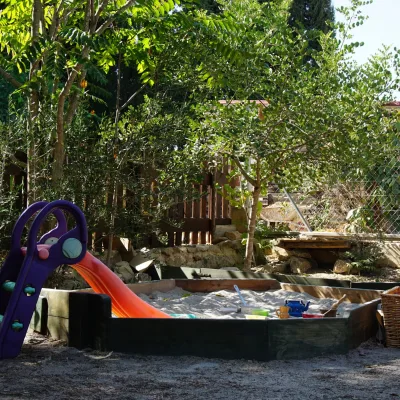 Arenero de madera con juguetes y tobogán infantil morado y naranja, rodeado de árboles y valla; área de juego al aire libre en un centro en la naturaleza.