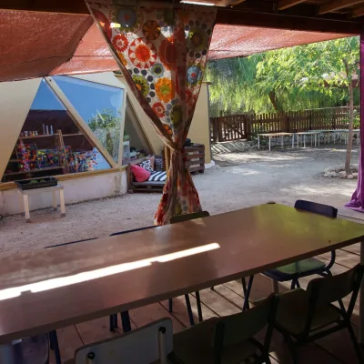 Mesas y sillas bajo porche con cortinas coloridas, frente a ventanales triangulares del domo y patio sombreado con árboles en un centro en la naturaleza.