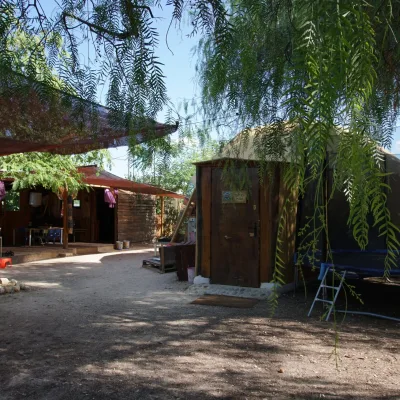 Zona exterior con mallas de sombra, edificaciones de madera y domo con puerta; árboles, camino de tierra y cama elástica en un centro en la naturaleza.