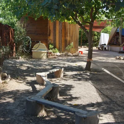 Pasarela de troncos para equilibrio bajo la sombra de un árbol con slackline lateral; al fondo fregadero y manguera junto a cabaña y domo, en un centro en la naturaleza.