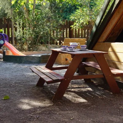 Mesa de picnic de madera con juego de té junto a cabaña infantil; al fondo, tobogán morado y arenero bajo los árboles en un centro en la naturaleza.