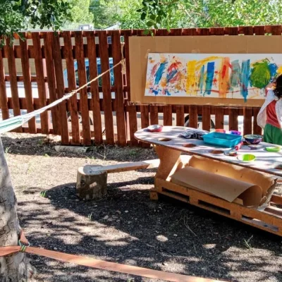 Niño pinta un gran mural en la valla junto a mesa de arte con temperas, mientras otro niño se columpia bajo un árbol; área exterior del centro en la naturaleza.